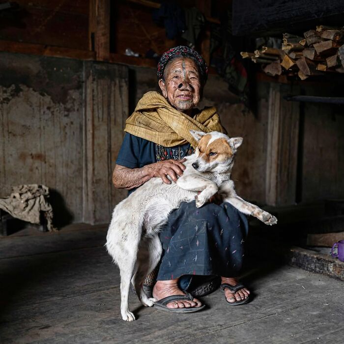 Elderly person sitting indoors, wearing traditional clothing, gently holding a dog in a striking portrait by Massimo Bietti.