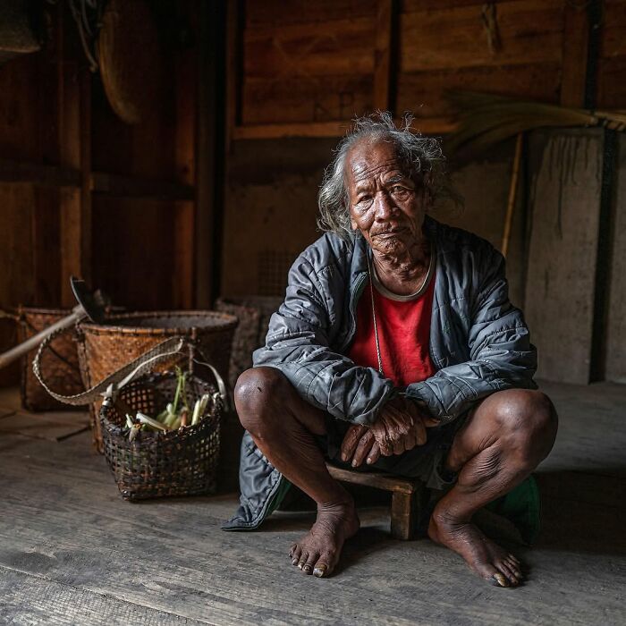 Elderly man sitting indoors by baskets of corn, captured in a striking portrait of elderly people across the globe.