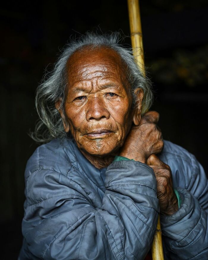 Elderly man with weathered face and gray hair holding a wooden stick in a striking portrait of elderly people.