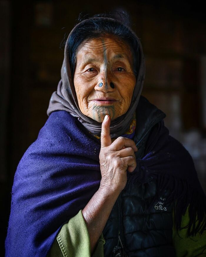 Elderly woman with traditional facial tattoos wearing a headscarf and purple shawl in a striking portrait by Massimo Bietti.
