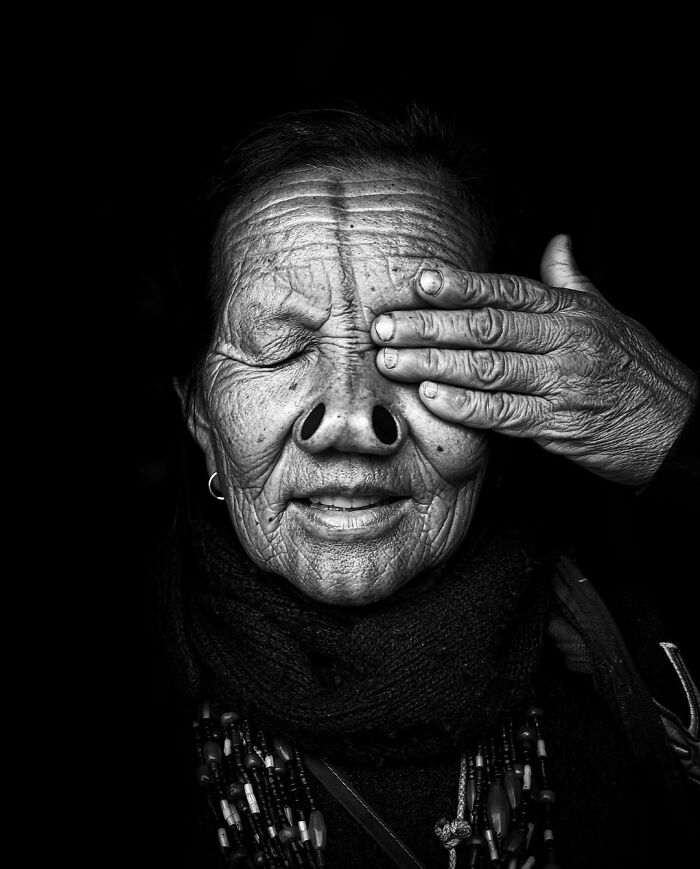 Black and white striking portrait of an elderly person with visible wrinkles and hand covering one eye in low light.