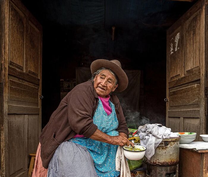 Elderly woman in traditional clothing preparing food inside a rustic home, captured in a striking portrait by Massimo Bietti.