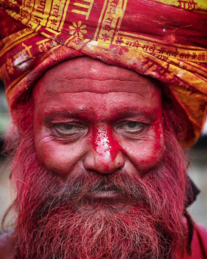 Elderly man with red-painted face and beard wearing a colorful traditional turban in a striking portrait.
