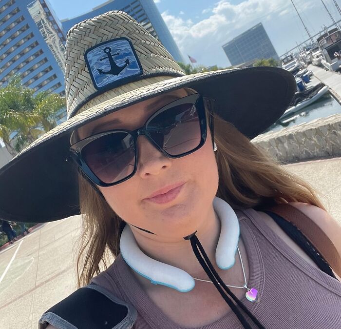 Woman wearing a wide-brimmed hat and sunglasses outdoors, staying cool with a neck fan, summer body game-changer accessory.