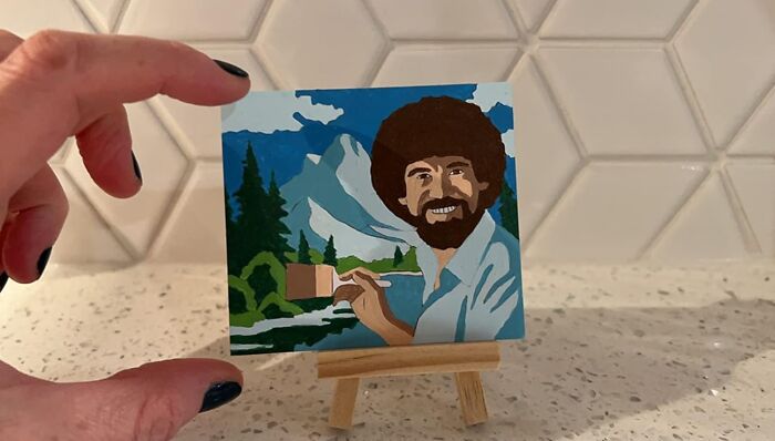 Hand holding a miniature Bob Ross painting on an easel, showcasing a fun hobby to trade screen time for skill time.