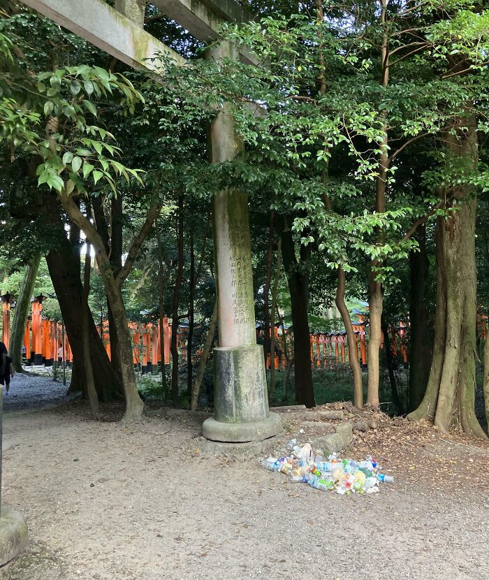 Pile of plastic bottles and trash left by entitled tourists in a forested area near a historic monument and pathway.