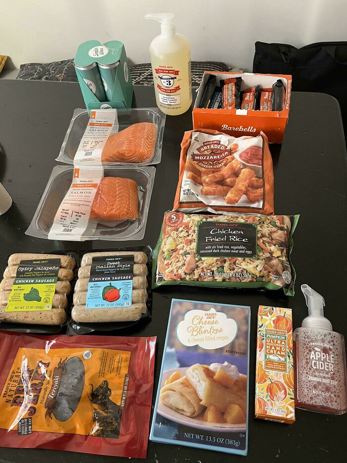 Groceries on a black table including salmon, sausages, frozen meals, snacks, and hand soap showing grocery cost.