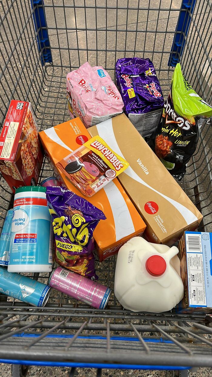 Shopping cart with various groceries including milk, soda, snack foods, and disinfecting wipes showing grocery cost differences.