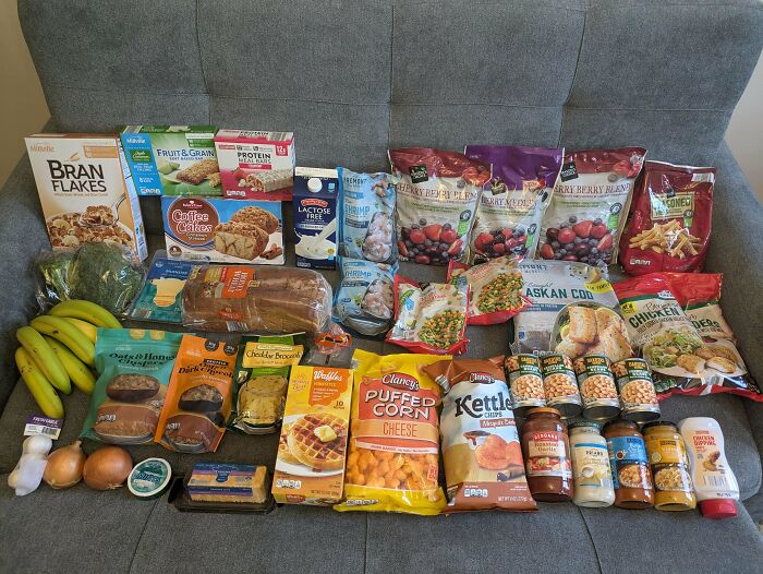Groceries spread on couch including fruits, snacks, frozen foods, dairy, and canned goods showing grocery costs where people live.