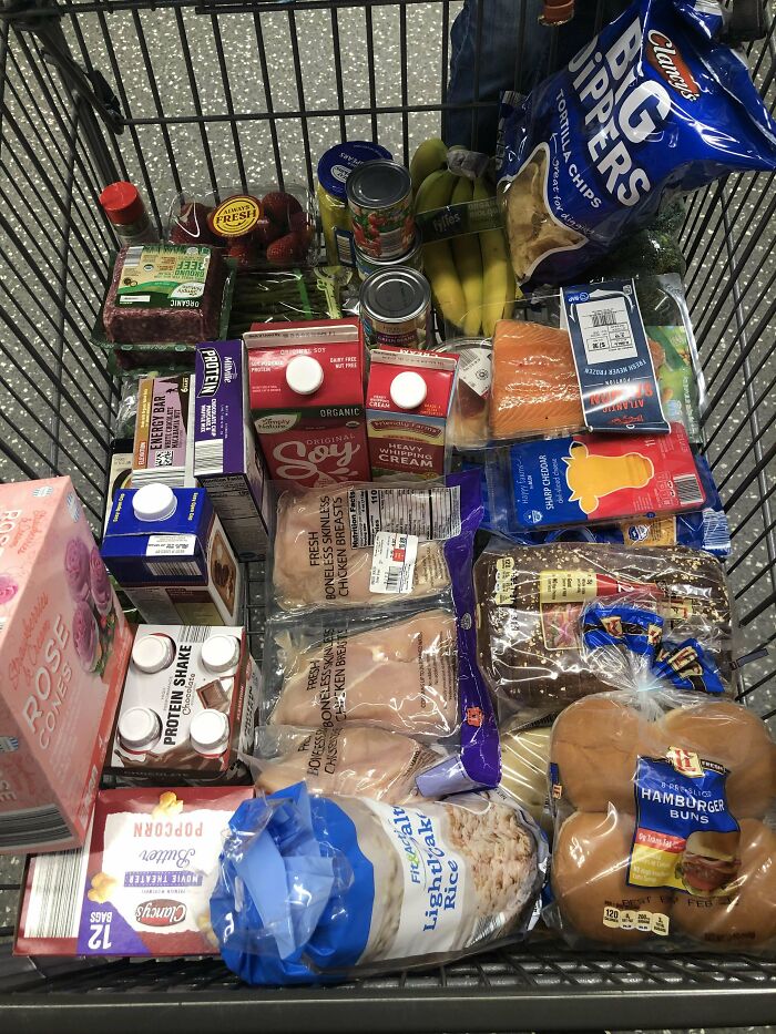 Grocery cart filled with various food items including produce, dairy, meat, snacks, and beverages showing grocery costs.