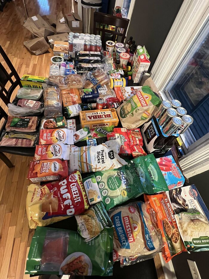 A large variety of groceries including snacks, dairy, meats, and drinks displayed on a dining table showing grocery cost differences.