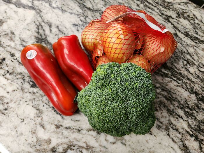 Red peppers, onions in a net bag, and broccoli on a marble countertop showing grocery cost items.