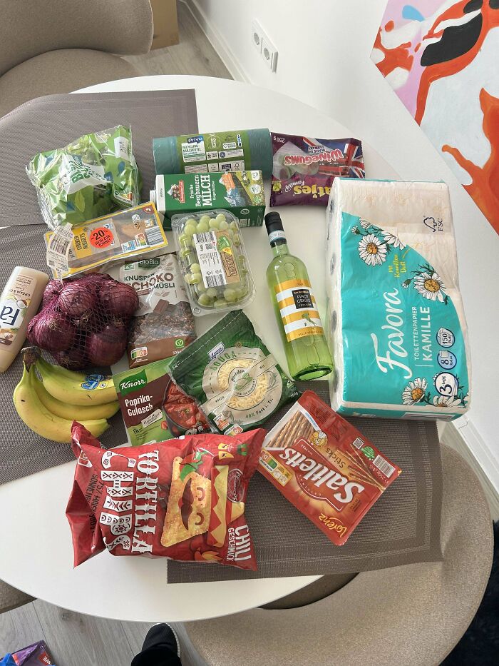 Groceries including fruit, snacks, toilet paper, and beverages displayed on a white table showing local grocery costs.