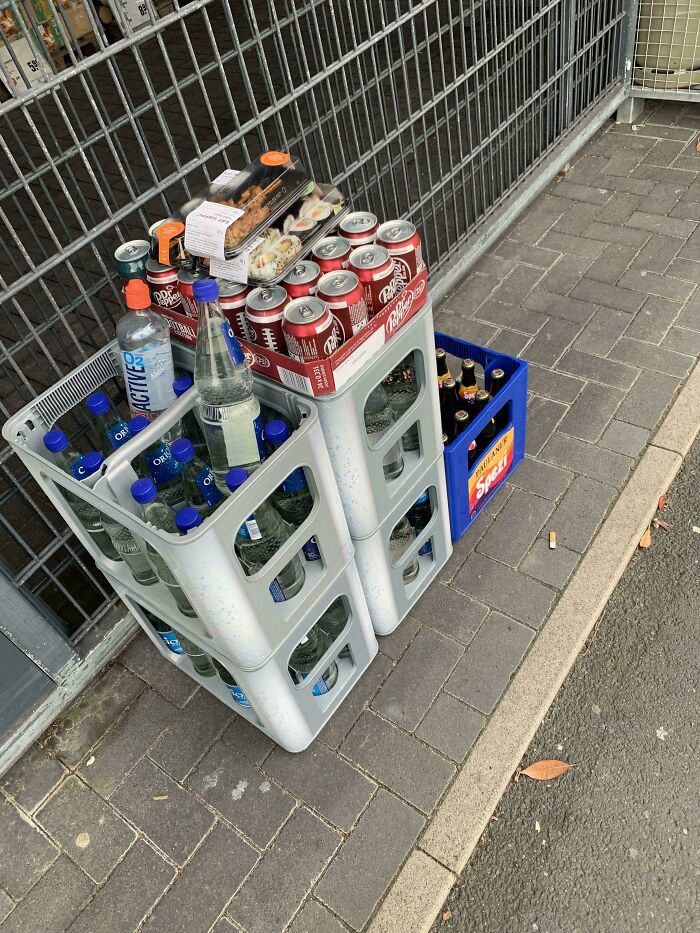Crates of bottled water, soda, and packaged sushi on a sidewalk showing grocery cost in different locations.