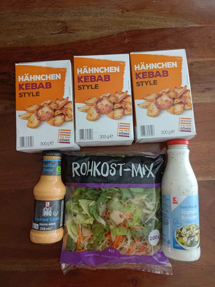 Three boxes of chicken kebab style, a bottle of cocktail sauce, a bag of salad mix, and a bottle of yogurt herb dressing on a wooden table.