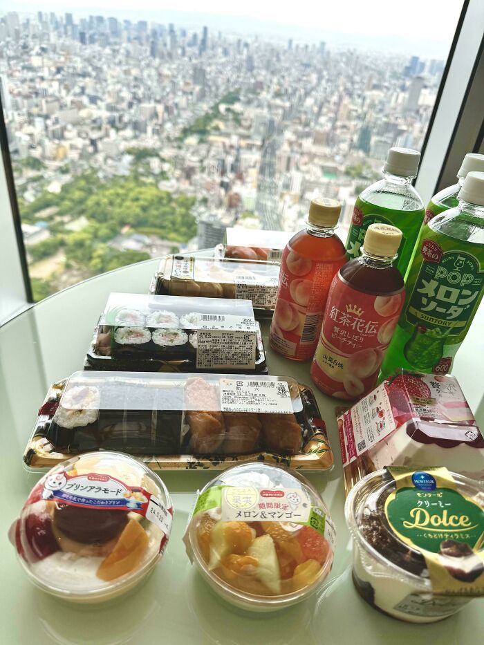 Variety of groceries including sushi, drinks, and desserts displayed on a table with a city view in the background.