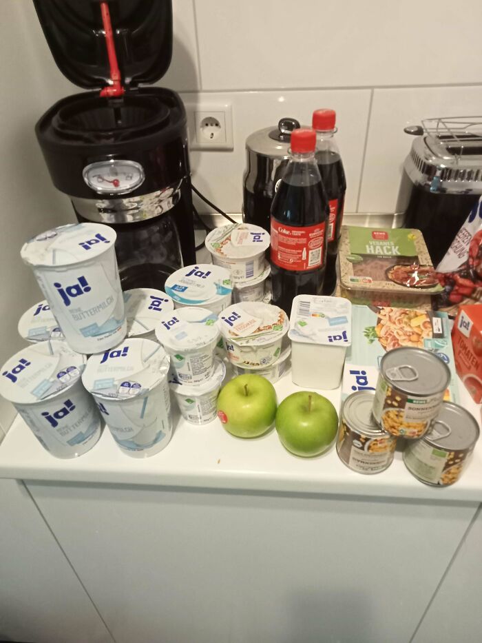 Groceries on a kitchen counter including yogurt, apples, soda, canned goods, and packaged food showing local grocery costs.