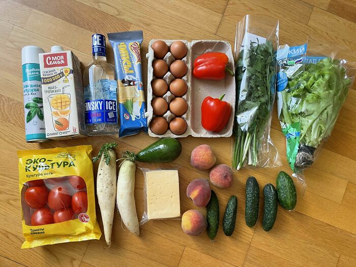 Grocery items including eggs, vegetables, peaches, cheese, and beverages arranged on a wooden floor showing grocery costs.