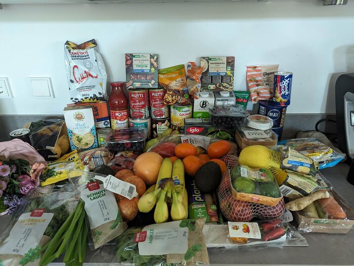 Groceries including fruits, vegetables, canned goods, and snacks displayed on a kitchen counter showing cost comparison.