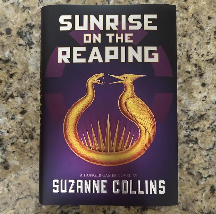 Book titled Sunrise on the Reaping by Suzanne Collins, with golden snake and bird forming a circle on purple background.