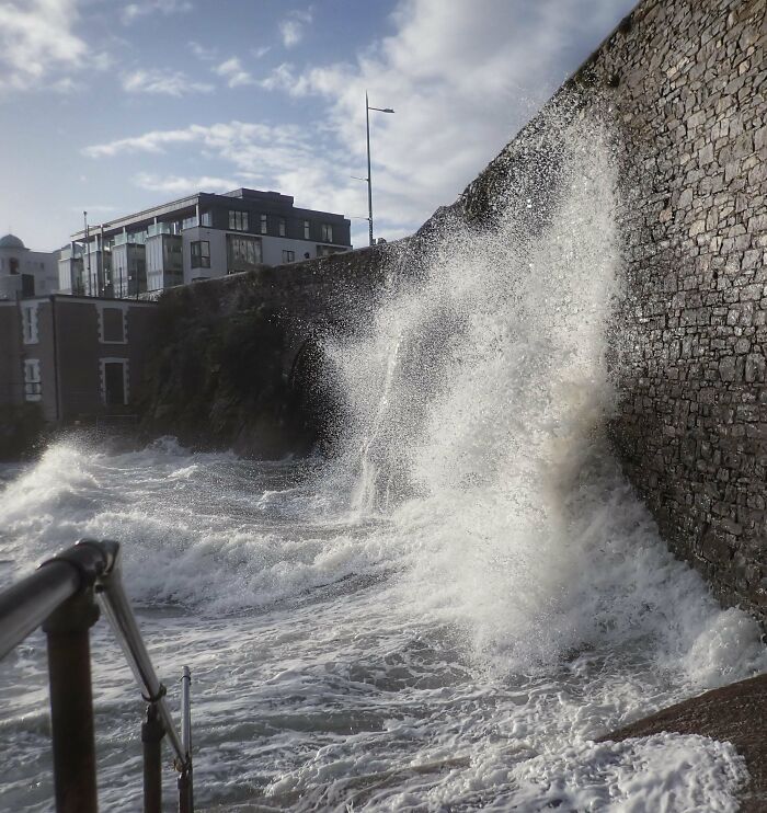 Waves crashing against a stone seawall with buildings in the background under a cloudy blue sky ocean creatures environment.