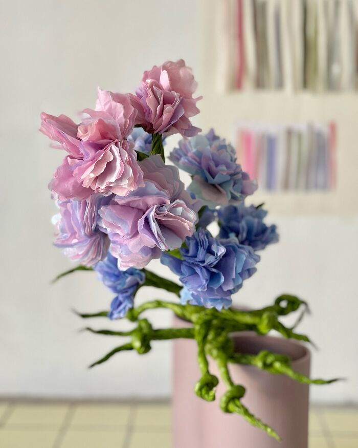 Giant flowers made of pastel tissue paper arranged in a pink vase with green twisted stems on a tiled floor.