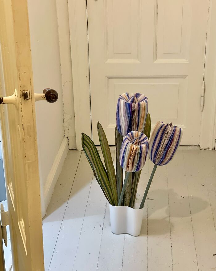 Giant flowers made from tissue paper with colorful petals and green leaves in a white container on a wooden floor.