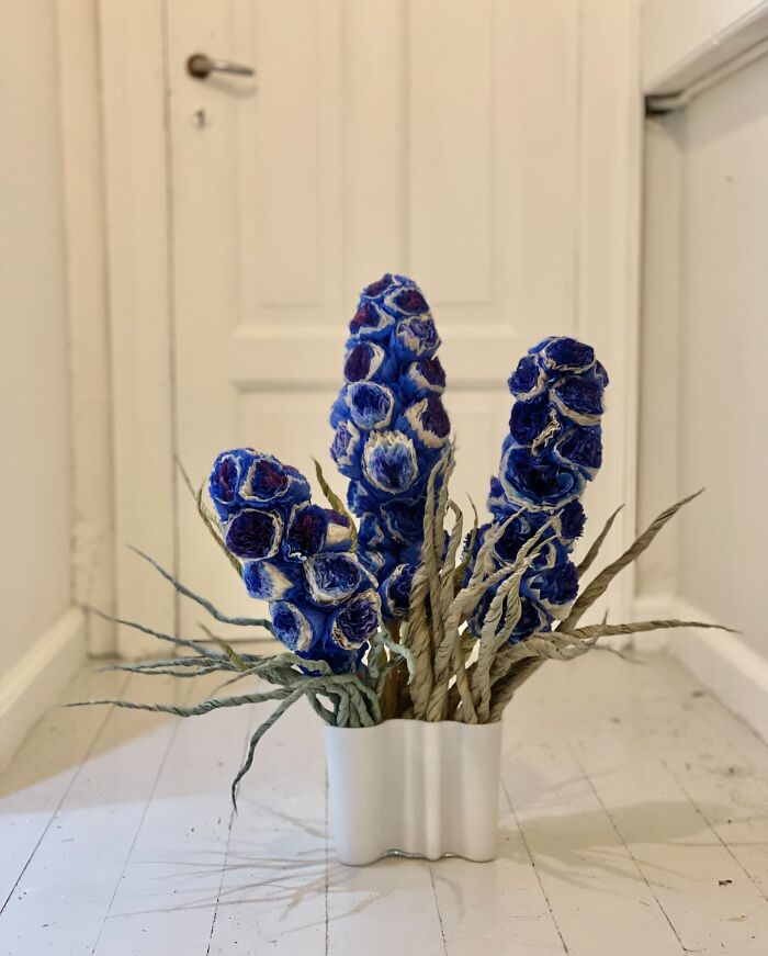 Giant flowers made using tissue paper in blue and purple shades arranged in a white vase on a wooden floor.