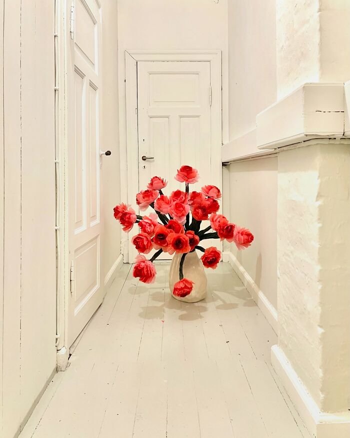 Giant tissue paper flowers arranged in a beige vase placed in a light-colored narrow hallway with white walls and doors.
