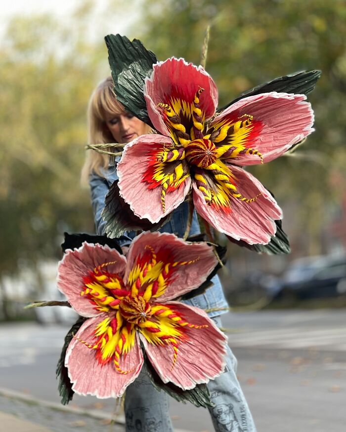 Woman holding giant tissue paper flowers with vibrant pink and yellow petals outdoors on a blurred background.