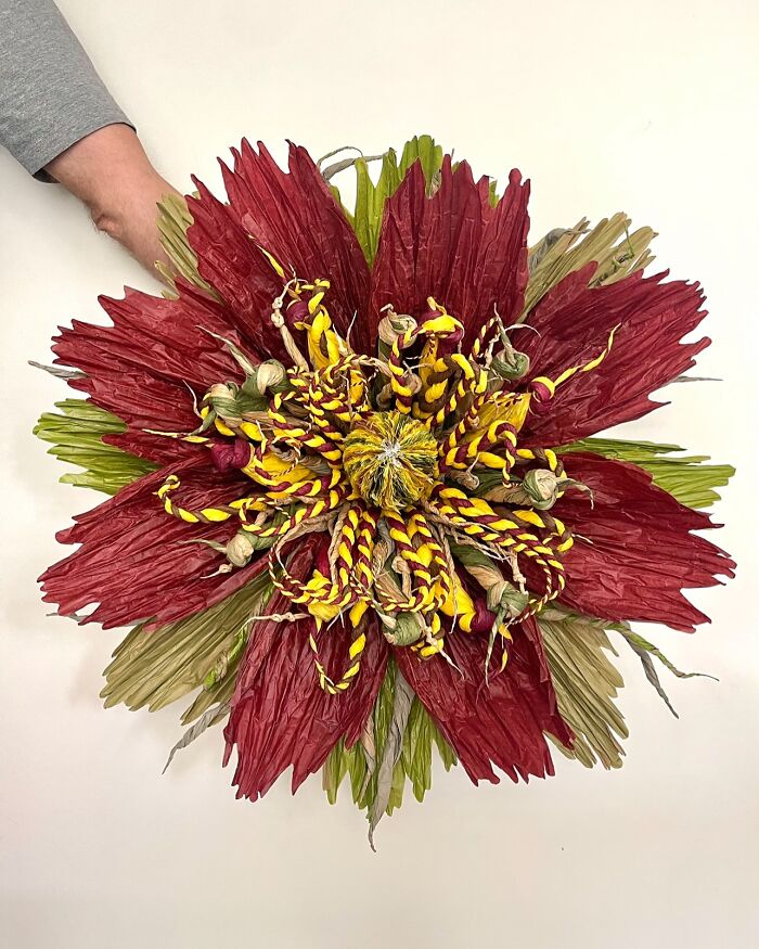 Giant tissue paper flower with red, yellow, and green petals held by a person wearing a gray shirt.