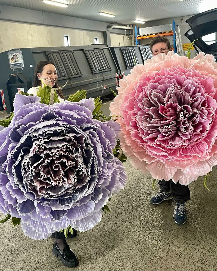 Two people holding giant tissue paper flowers, showcasing large, detailed, colorful handmade floral art.