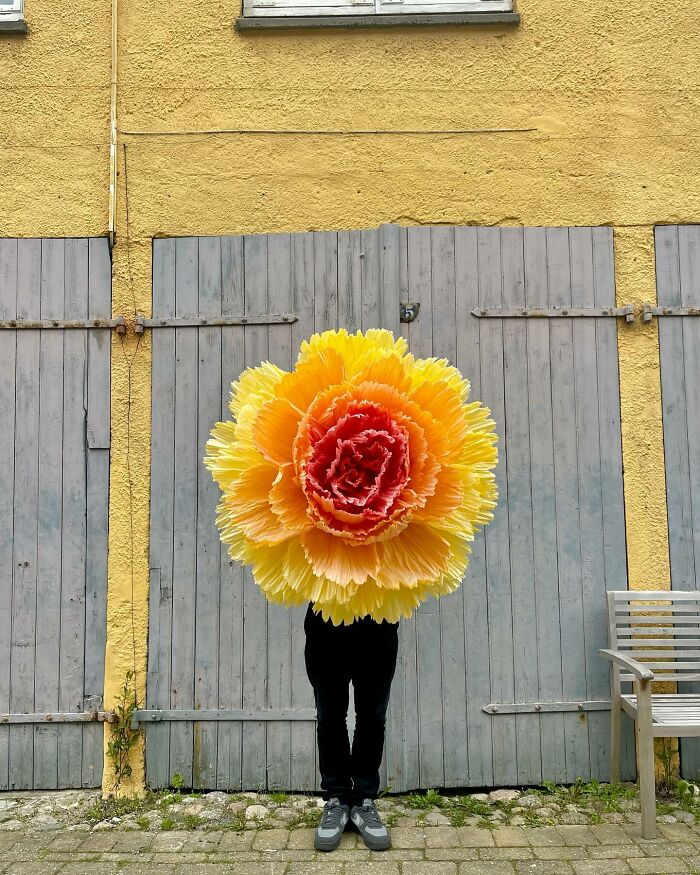 Person holding a giant tissue paper flower with yellow and red petals standing against a yellow wall and gray doors.