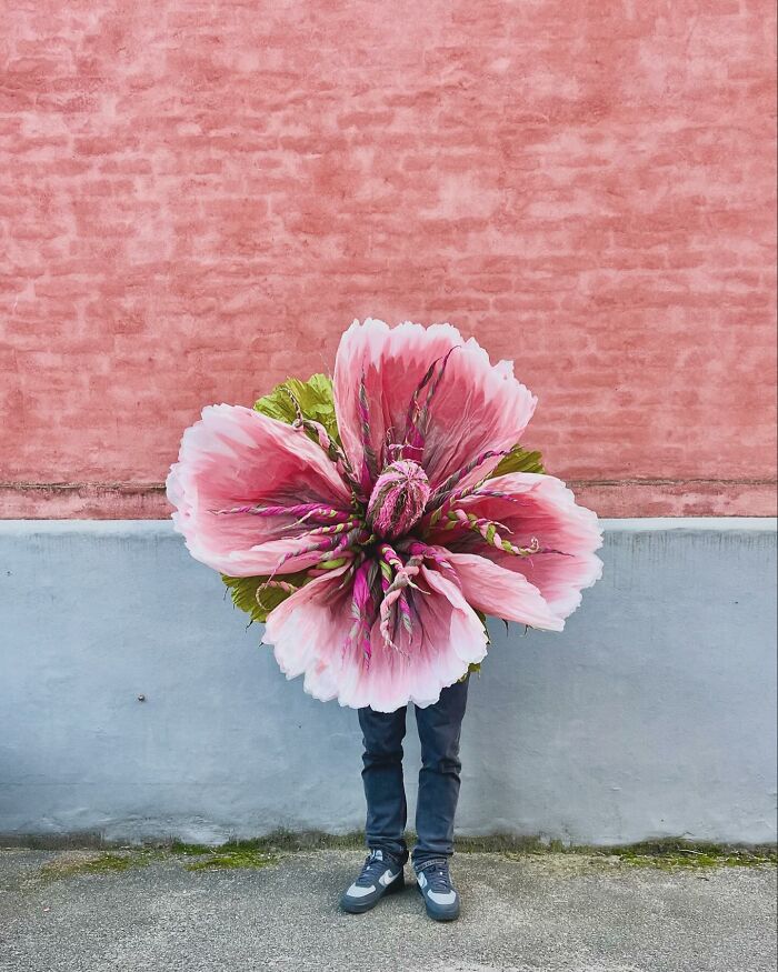 Person holding a giant tissue paper flower with pink petals standing against a half pink, half gray wall background.