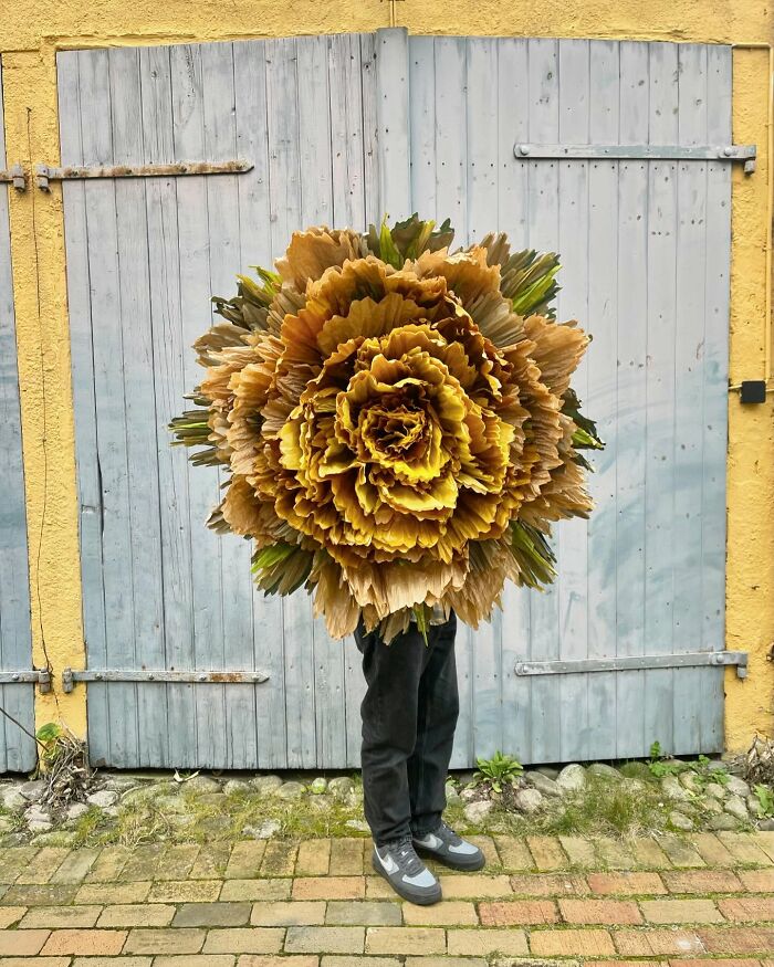 Person holding a giant flower made of tissue paper with layered yellow and green petals in front of wooden doors.