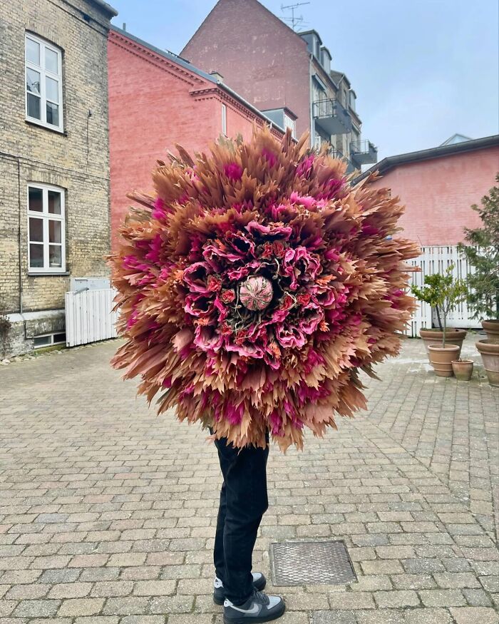 Woman holding a giant flower made of tissue paper in an outdoor urban setting with brick buildings.