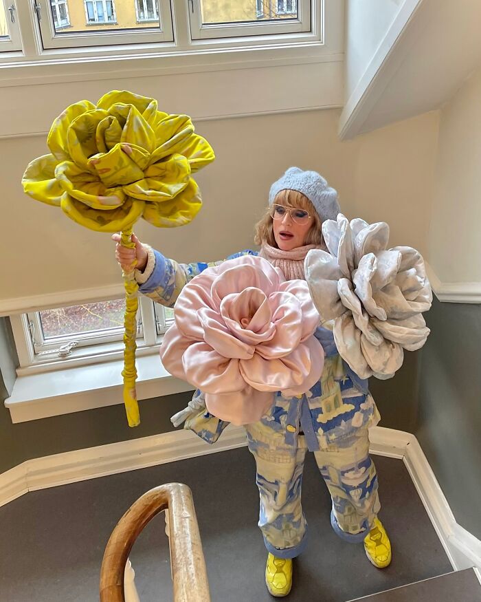 Woman holding giant tissue paper flowers in yellow, pink, and white, showcasing her latest oversized floral creations indoors.