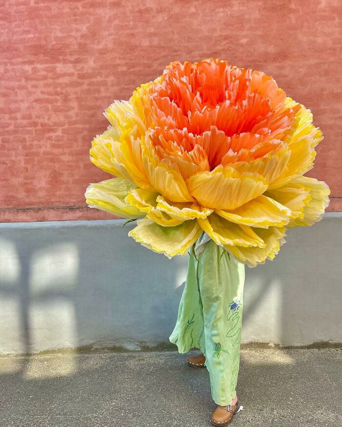 Giant tissue paper flower in bright yellow and orange hues held by a person in green pants and brown shoes outdoors.