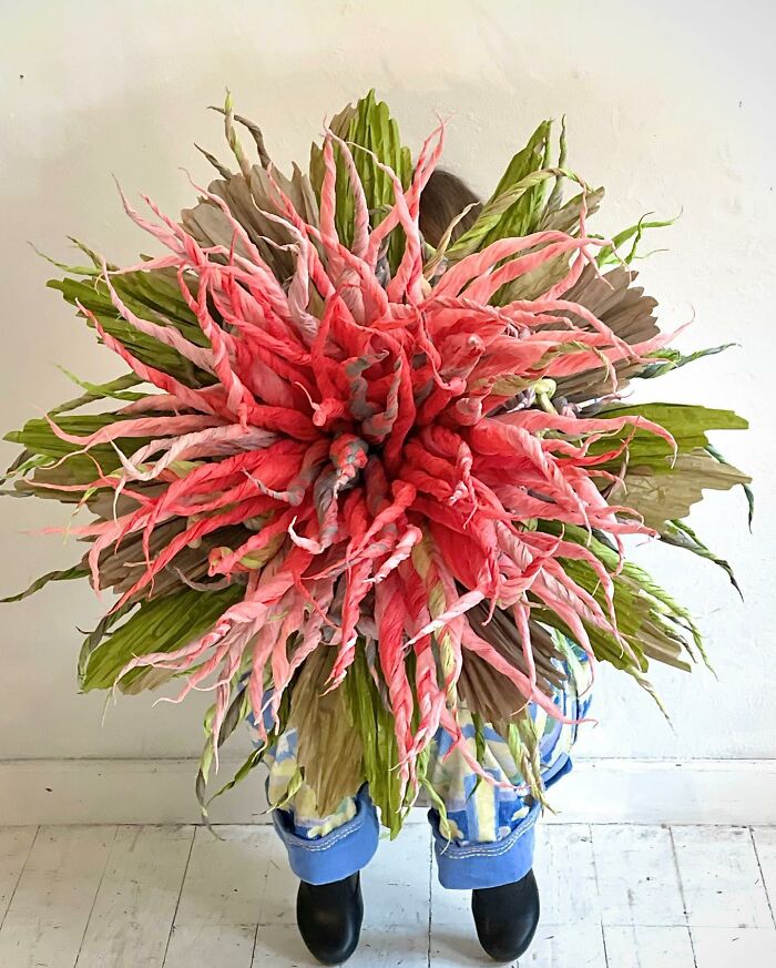 Giant flower made of tissue paper with pink twisted petals and green leaves held by a person wearing blue jeans and boots.