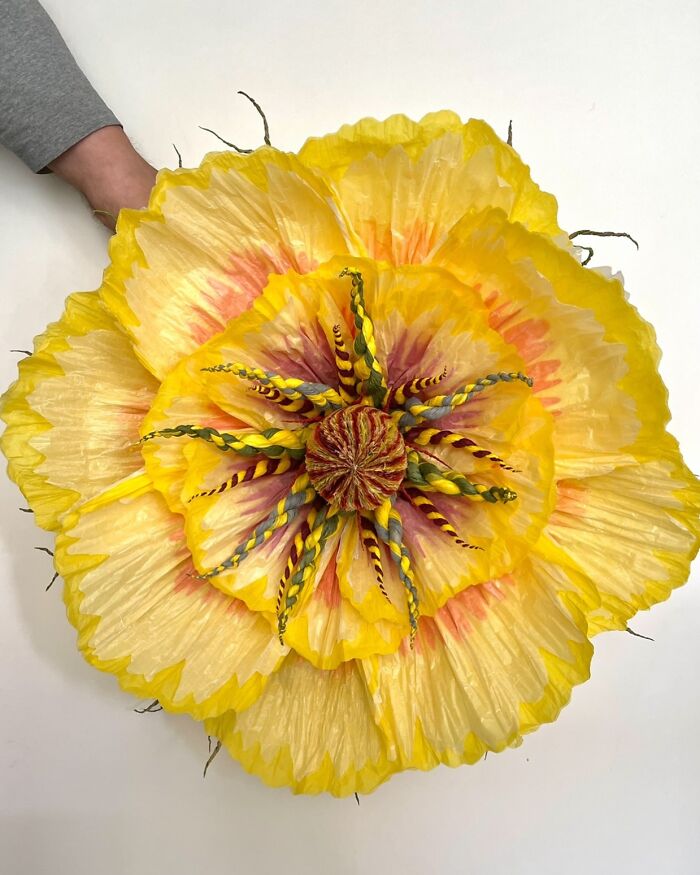 Hand holding a giant tissue paper flower with yellow and orange petals and intricate twisted center details.