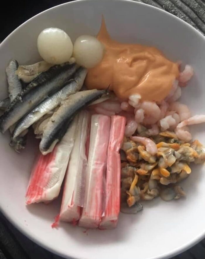 Bowl of mixed seafood including small fish, shrimp, crab sticks, clams, onions, and orange sauce showing cursed food.