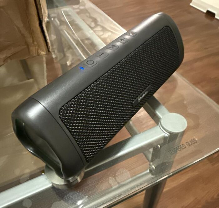 Portable Bluetooth speaker with control buttons, a blue indicator light, and mesh speaker grille on a glass table, summer essentials.