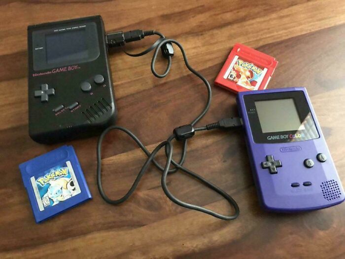 Two vintage Game Boy devices connected by a link cable with classic Pokémon game cartridges on a wooden surface.