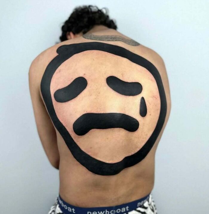 Large black sad face tattoo covering most of a person's back, showcasing funny and interesting bad tattoos.