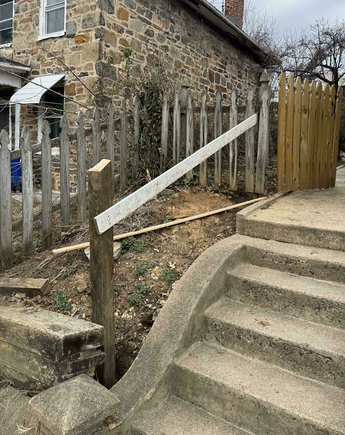 Improvised broken wooden handrail by stairs next to old stone house and weathered picket fence, landlord special fail.