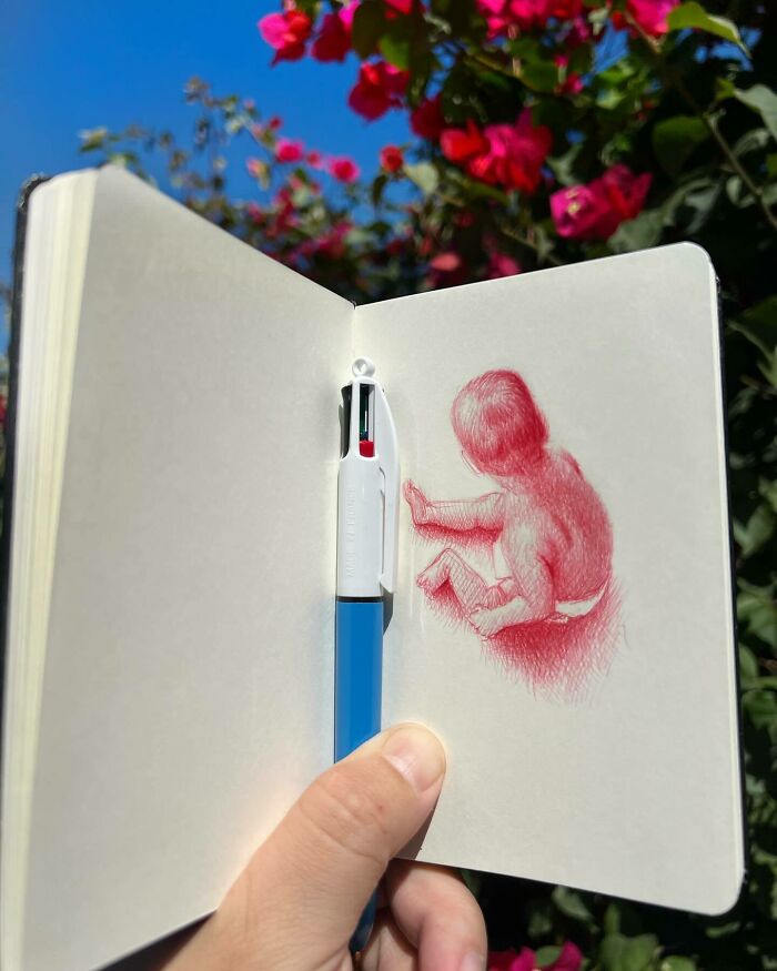 Tiny sketchbook held open outdoors showing a red ballpoint drawing of a child, capturing big emotions in everyday moments.