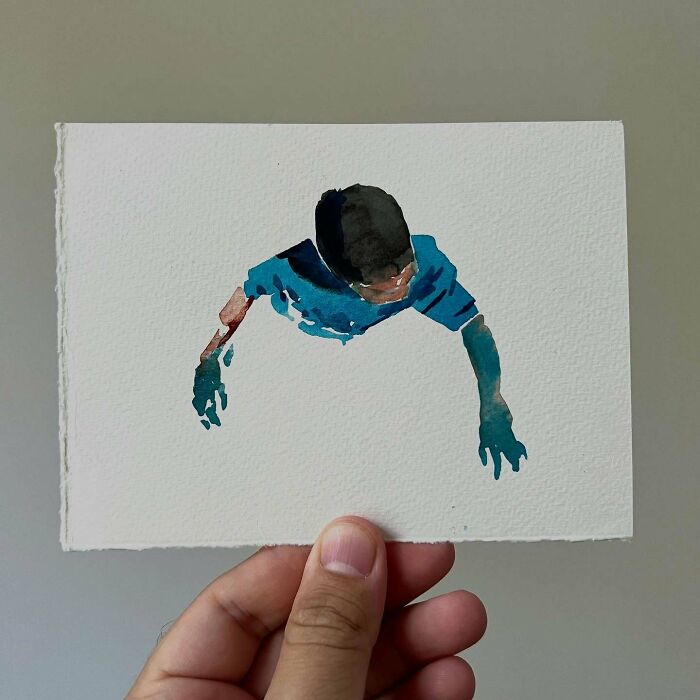 Hand holding a tiny sketchbook with a watercolor painting of a person from a top-down view, showing big emotions.