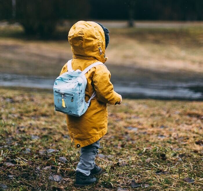 Child in a yellow coat and blue backpack standing outdoors, symbolizing dark family secrets and childhood reflections.