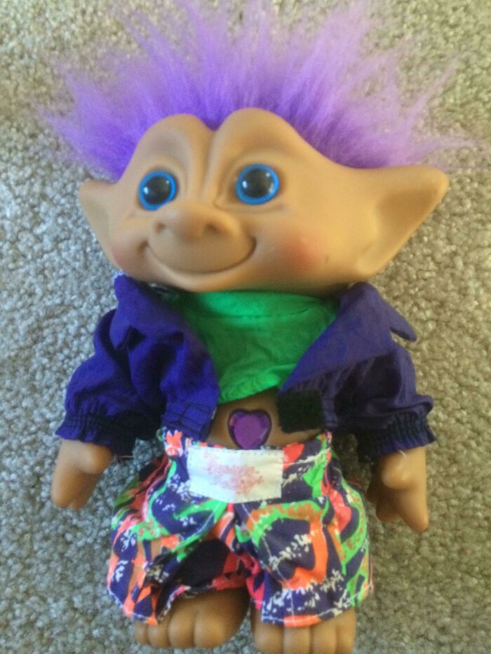 Troll doll with bright purple hair wearing cringey nineties fashions colorful shorts and a purple jacket on carpet background.