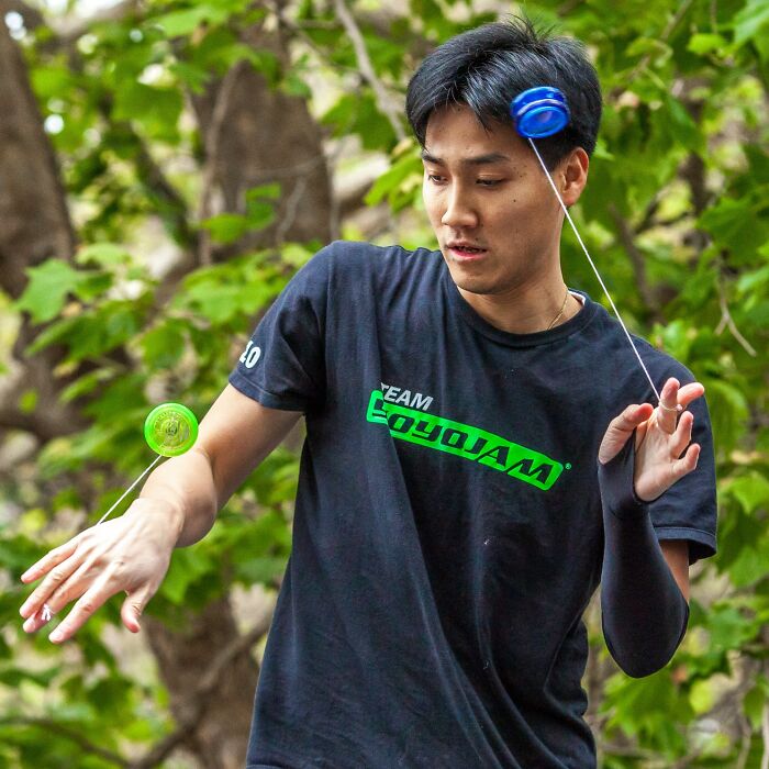 Young man outdoors wearing a cringey nineties fashion black shirt, playing with a blue and green yo-yo among green trees.